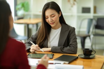 Asian woman writing on ipad at a desk with another person